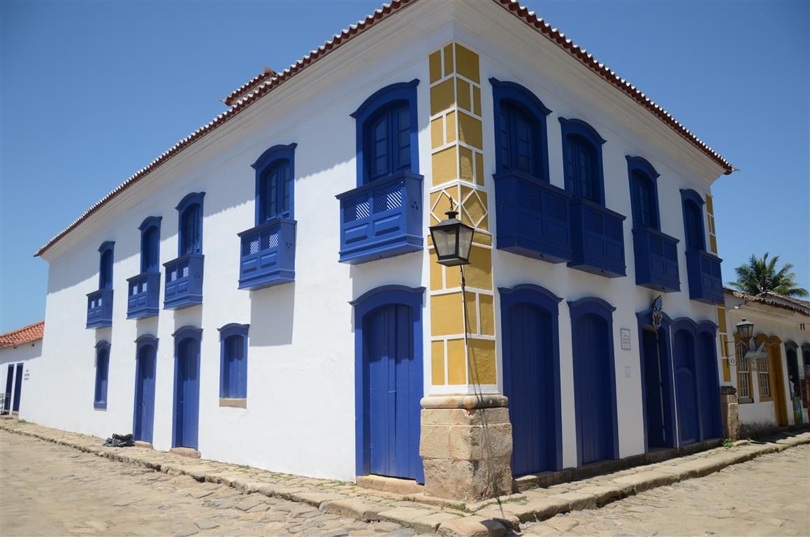 Prédio branco de dois andares com venezianas azuis e varandas em uma rua ensolarada de paralelepípedos.