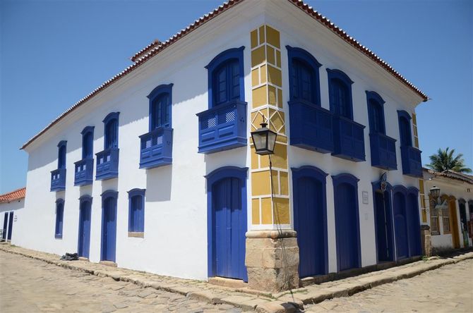 Prédio branco de dois andares com venezianas azuis e varandas em uma rua ensolarada de paralelepípedos.
