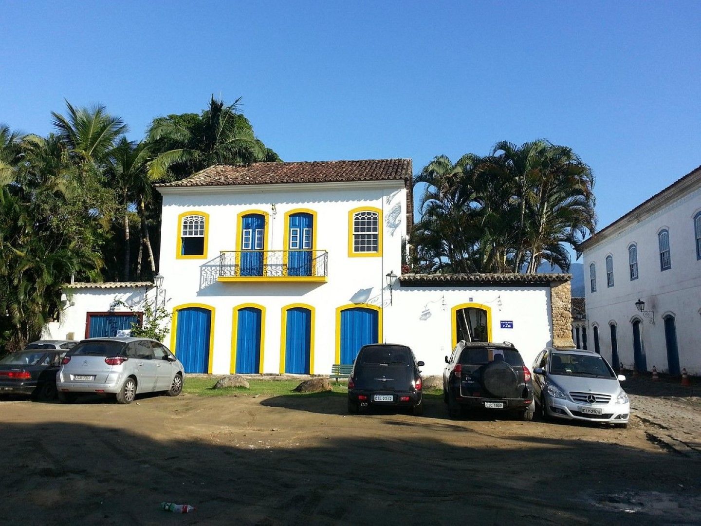 Um edifício colonial branco com portas azuis e detalhes amarelos em um ambiente externo ensolarado; carros estacionados na frente.