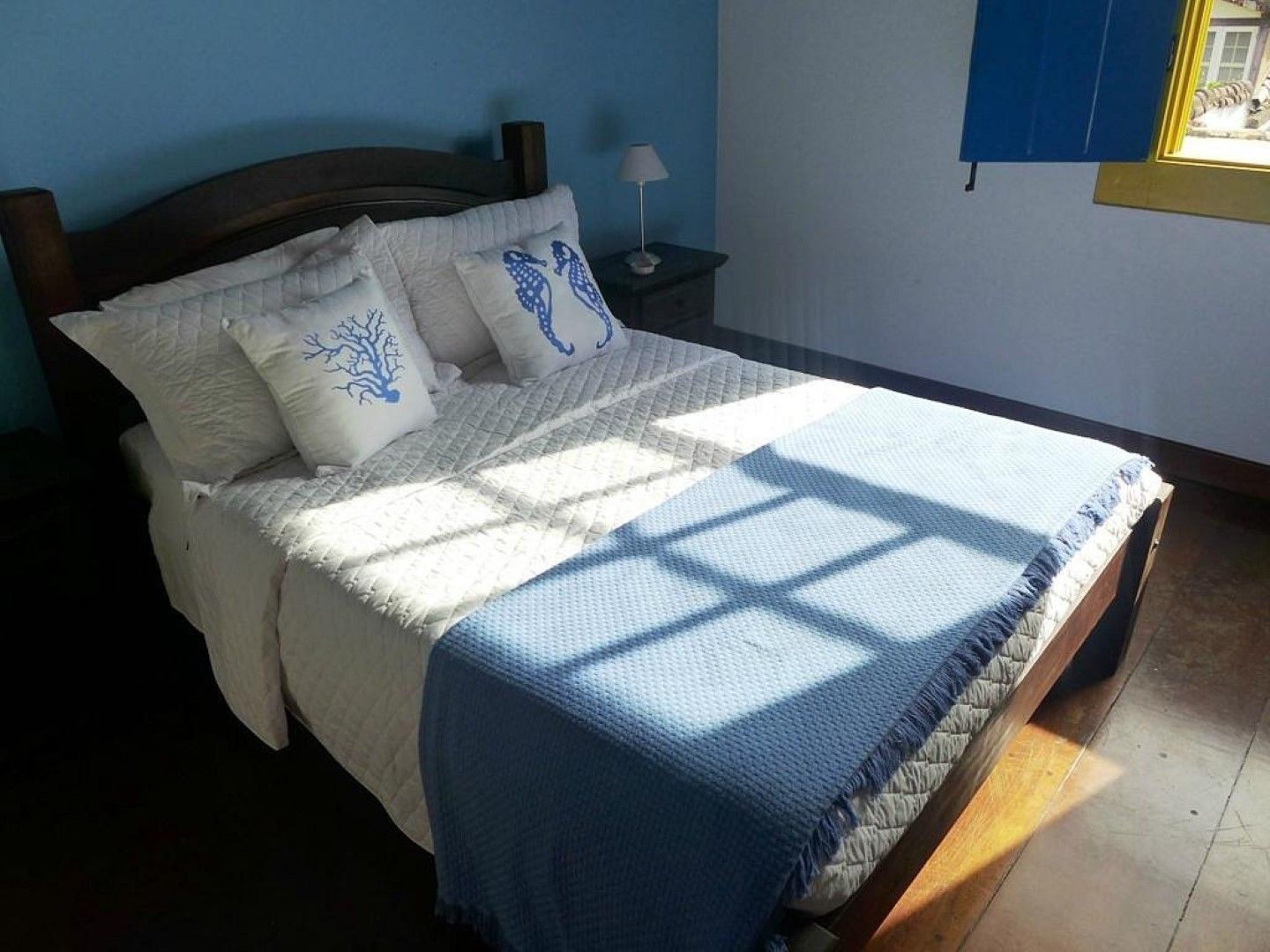 Quarto com cama coberta com roupa de cama branca, manta azul e almofadas decorativas. A luz do sol entra pela janela, iluminando o ambiente.