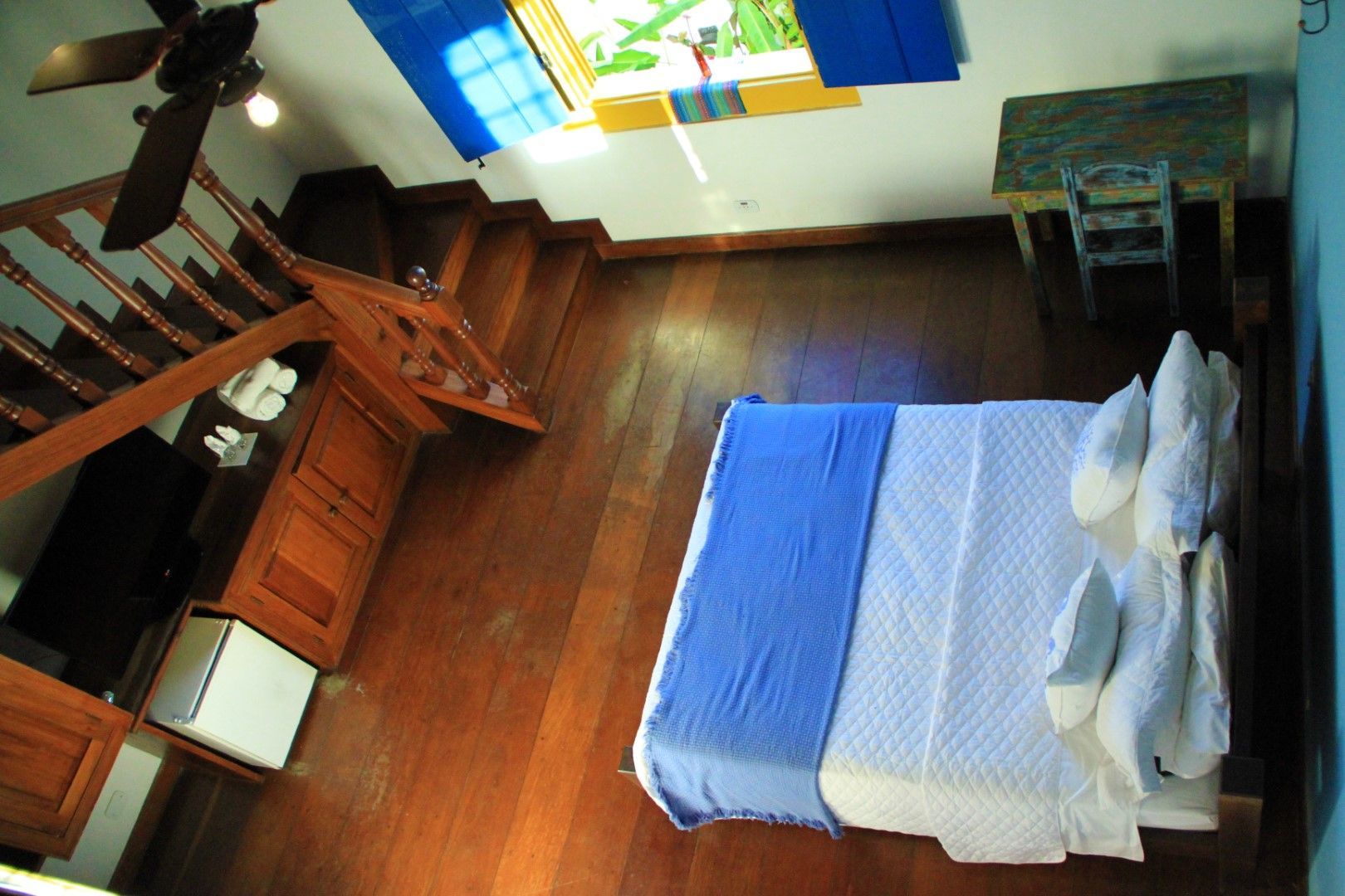 Quarto com piso de madeira, escadas, cama com cobertas azuis, uma escrivaninha e uma claraboia.
