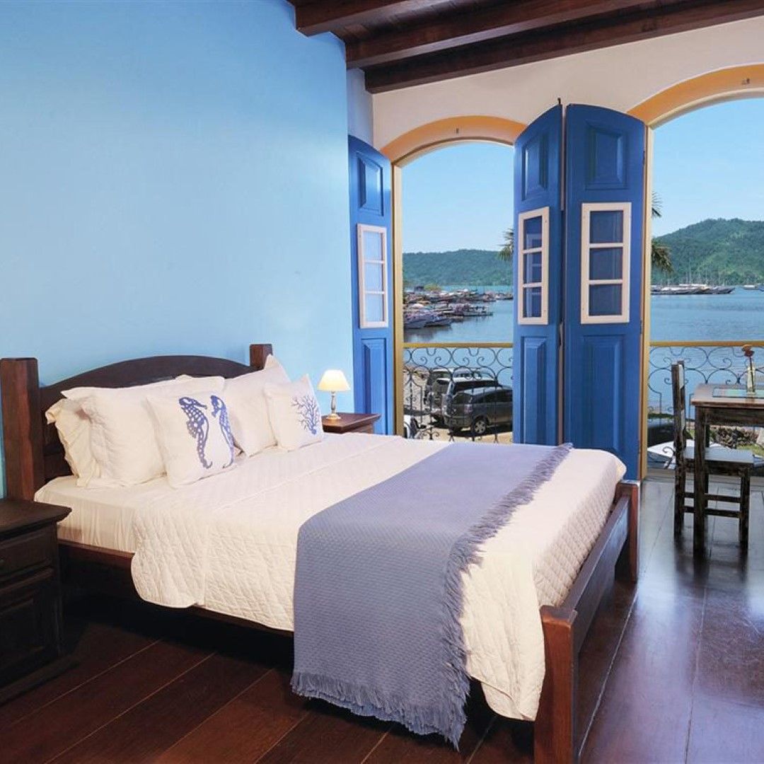 Quarto com cama, paredes azuis, móveis de madeira e varanda com vista para uma baía com barcos.
