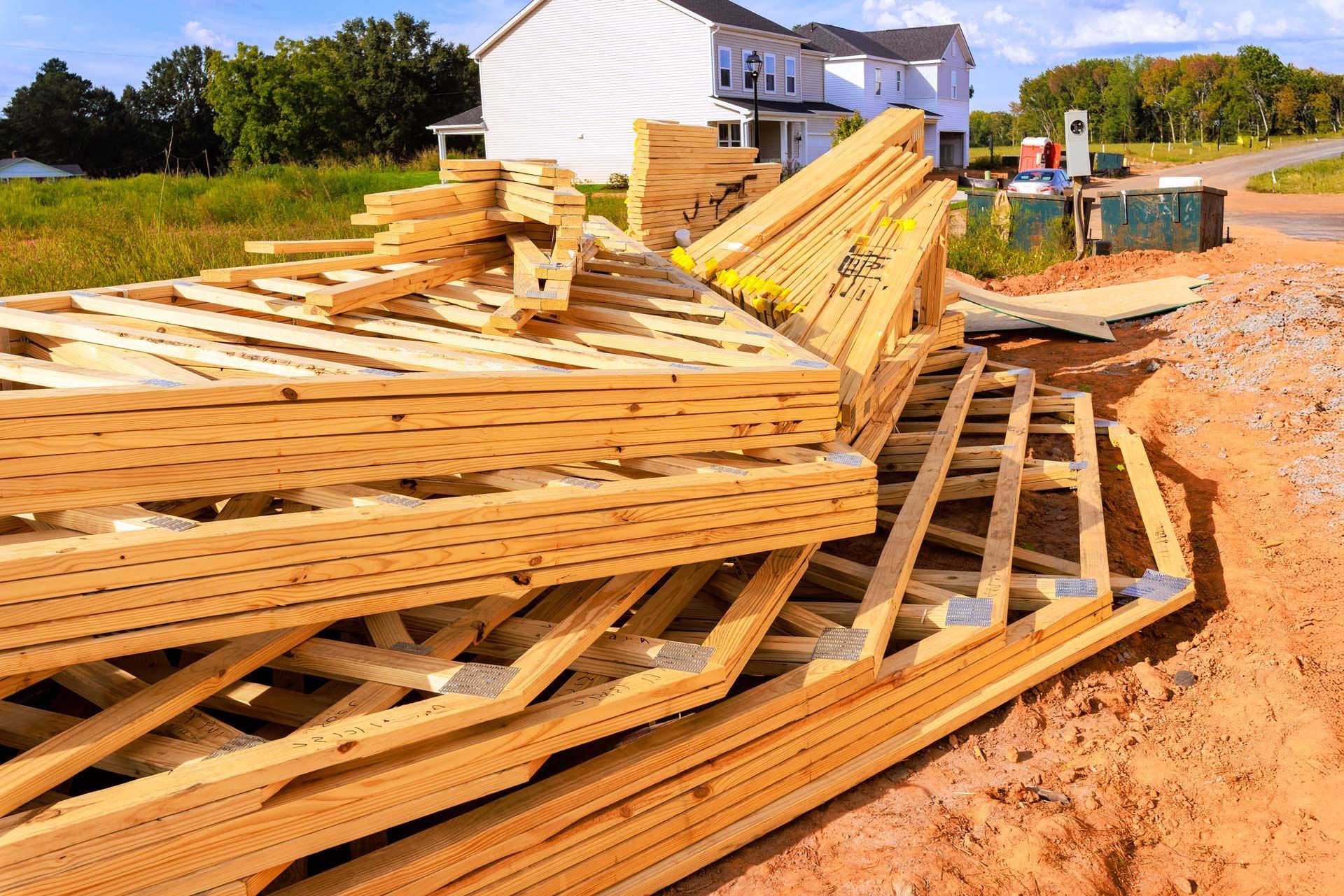 Stack of wooden roof trusses at a residential construction site with a house in the background.