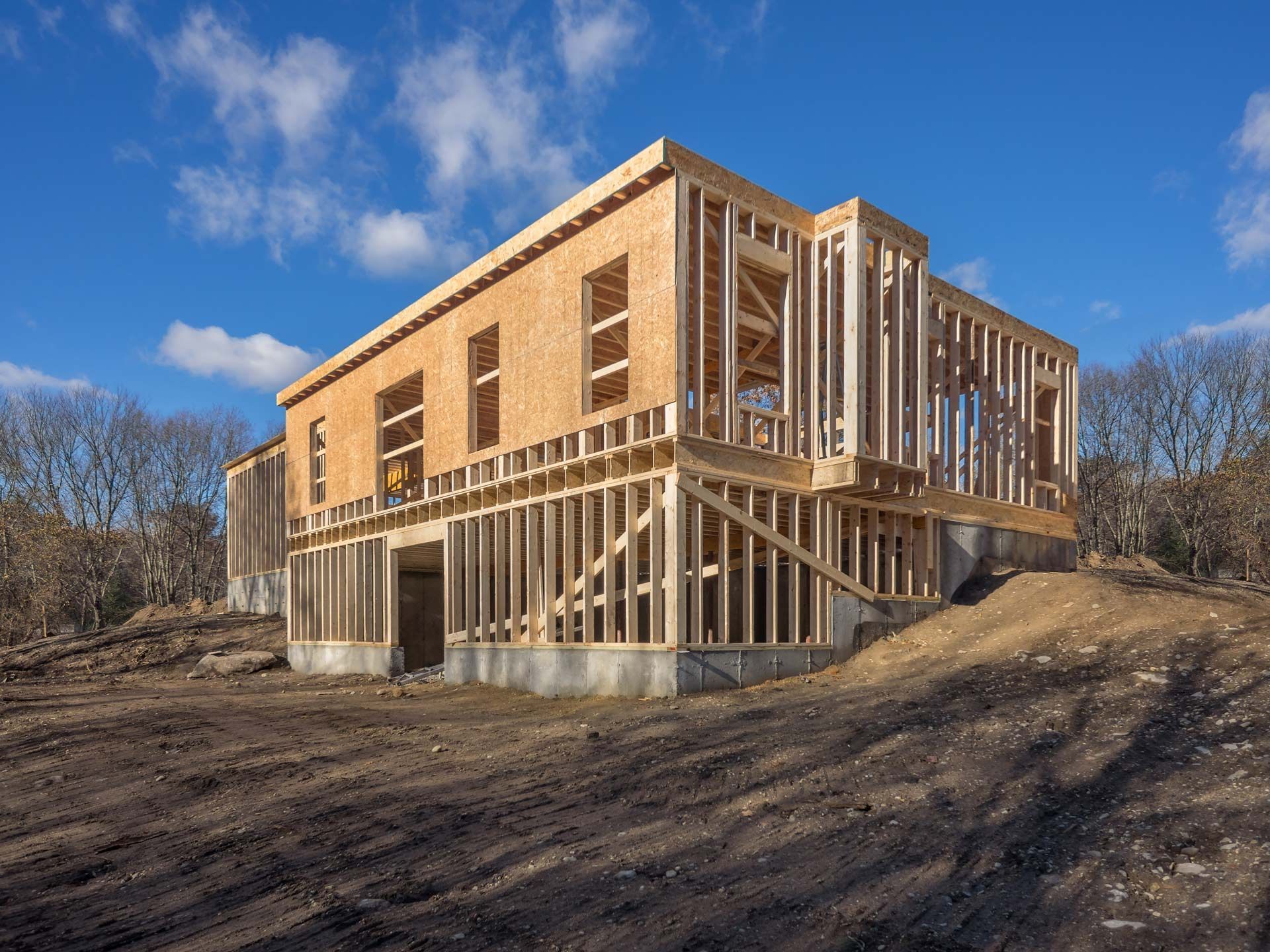 The wooden frame of a two-story house under construction stands on a dirt lot against a bright blue sky.