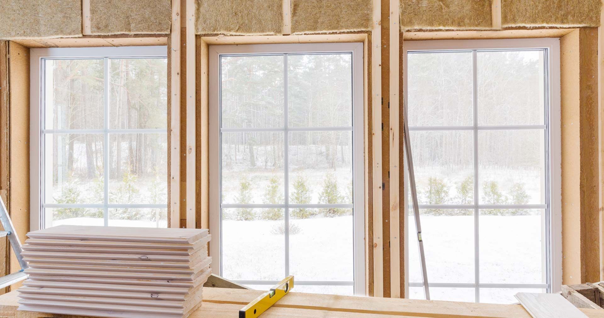 Three white-framed windows set into a wooden wall frame under construction, with drywall stacks in the foreground.
