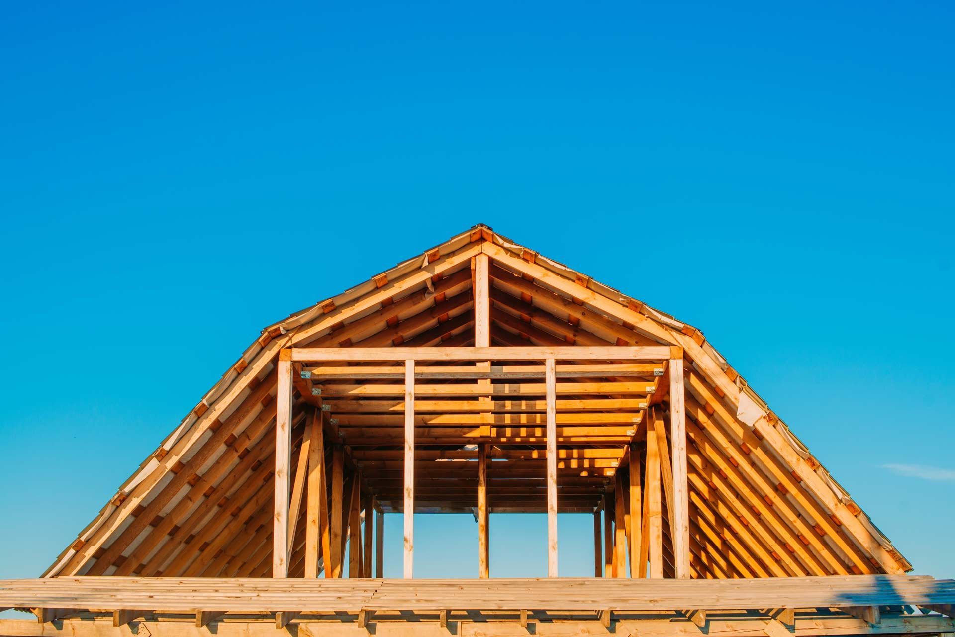 The wooden frame of a house roof construction stands against a clear, bright blue sky.