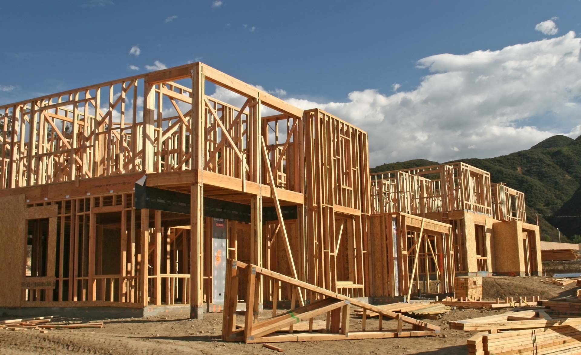 Wooden frames of several unfinished houses under construction against a mountain backdrop under a blue sky.
