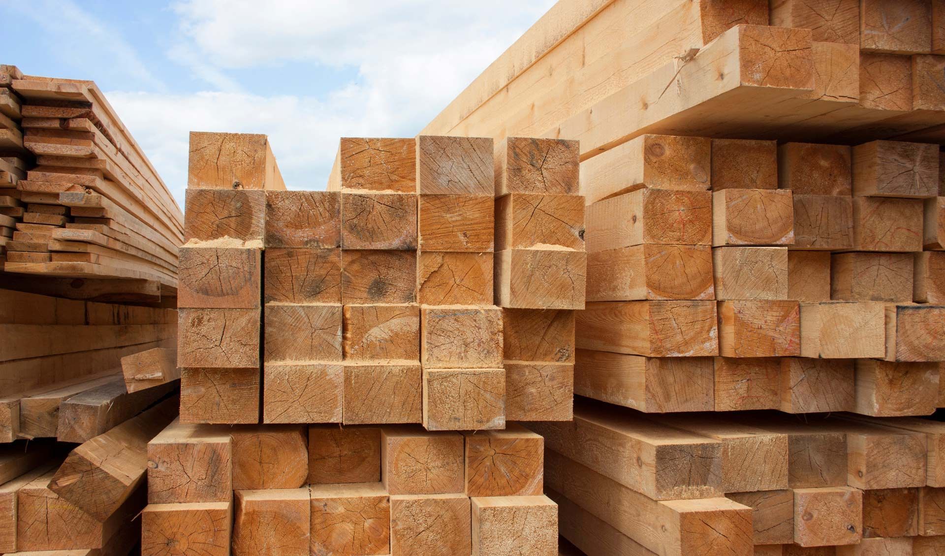 Stacks of light brown lumber beams piled outdoors against a cloudy blue sky.