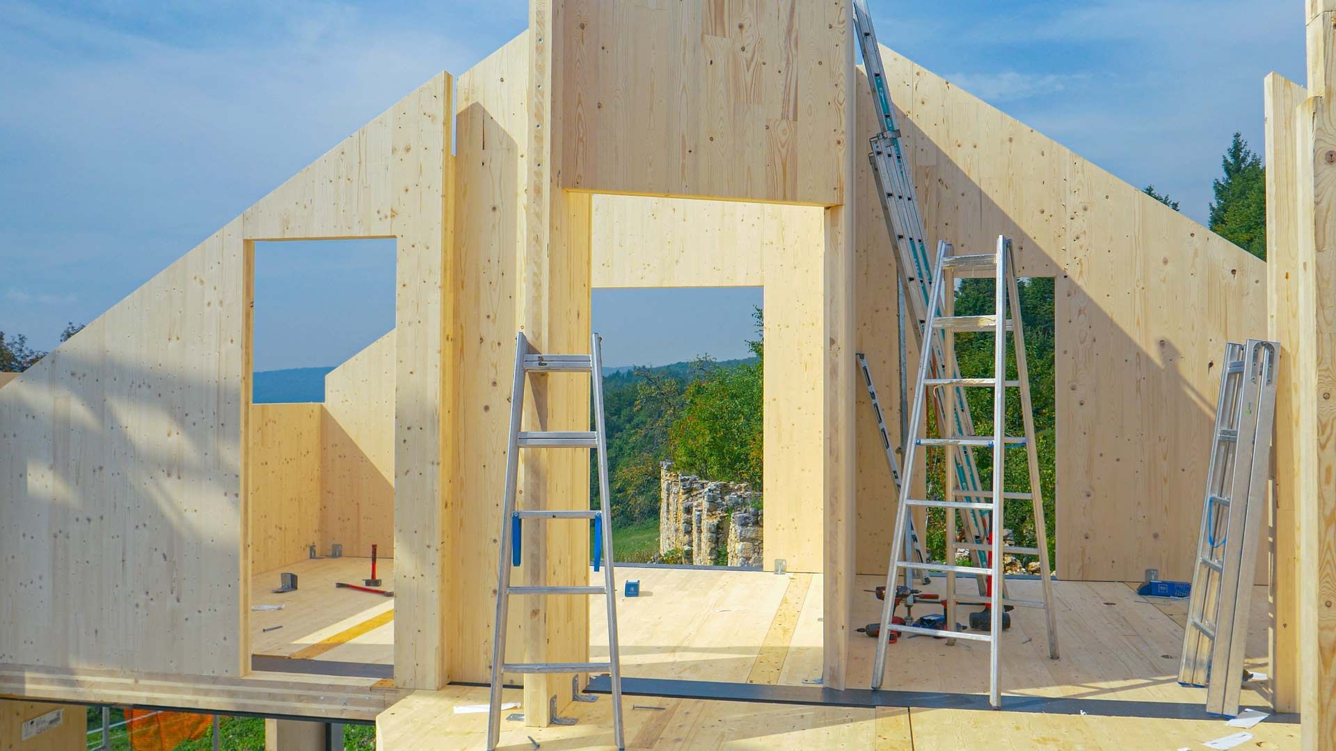 Construction site with light wood panels for walls, door frames, and multiple metal ladders under a bright blue sky.
