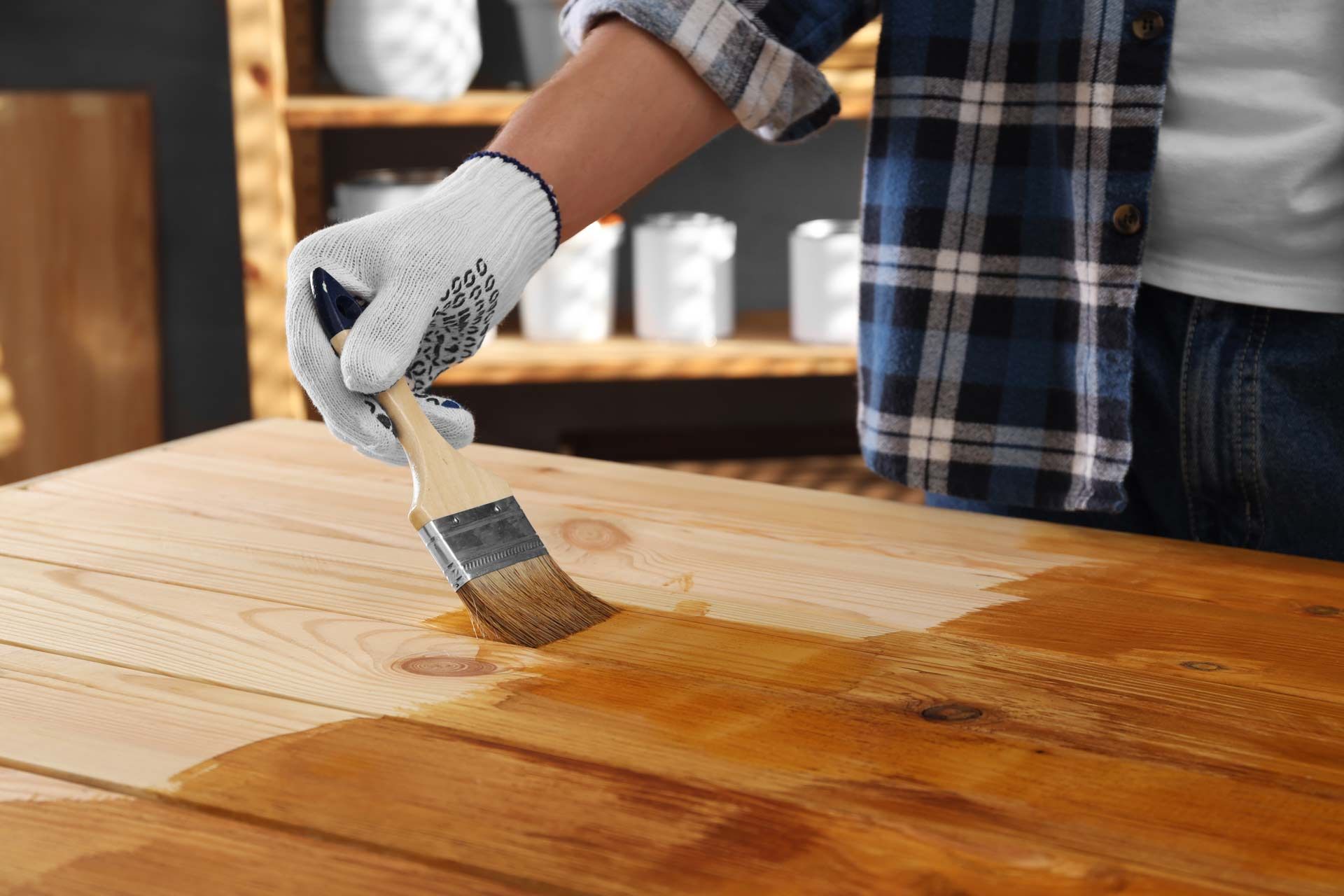 A person wearing a glove uses a brush to apply a brown wood stain to a light-colored wooden table.