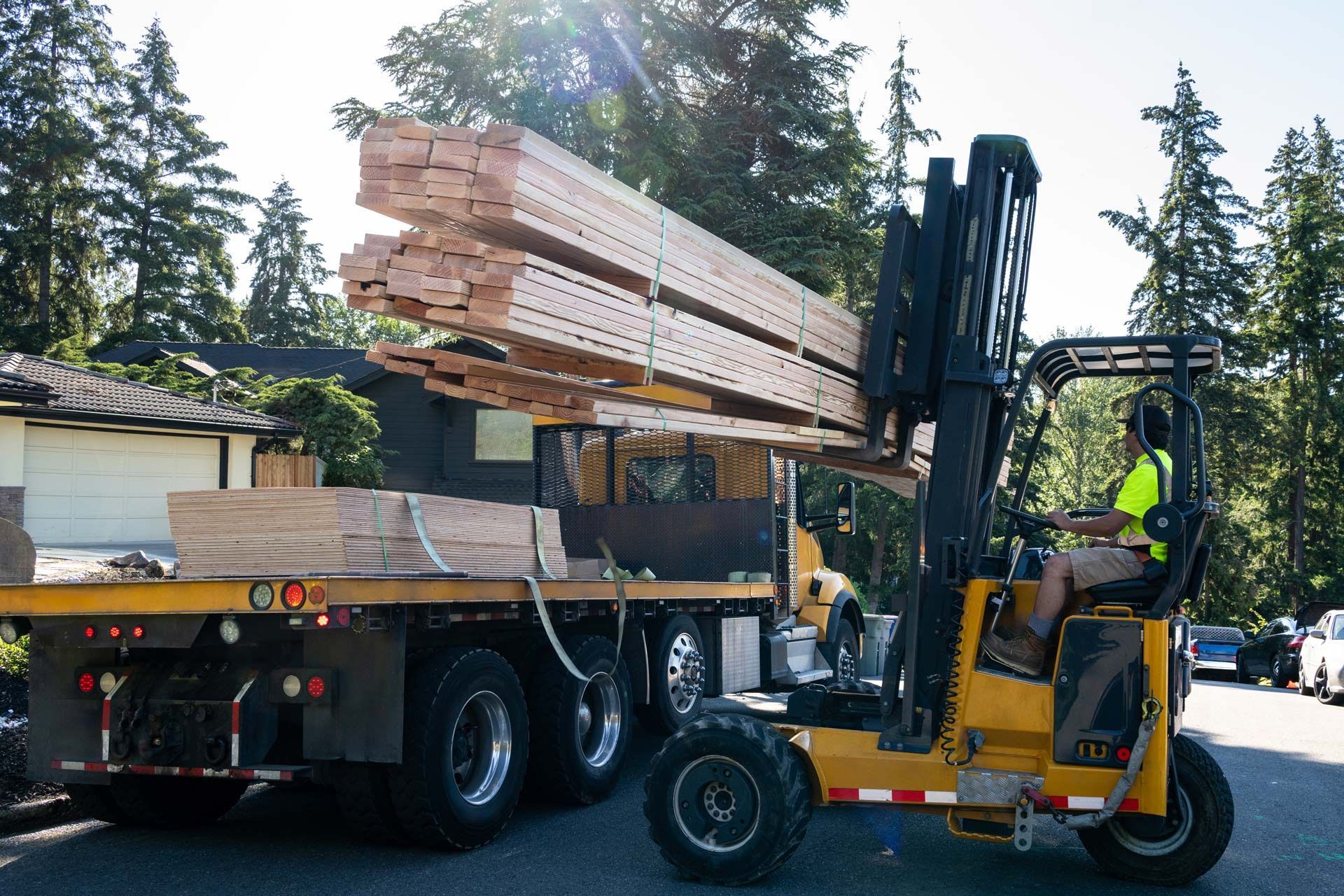 A yellow forklift lifts a large bundle of lumber from a flatbed truck on a sunny day near residential trees.