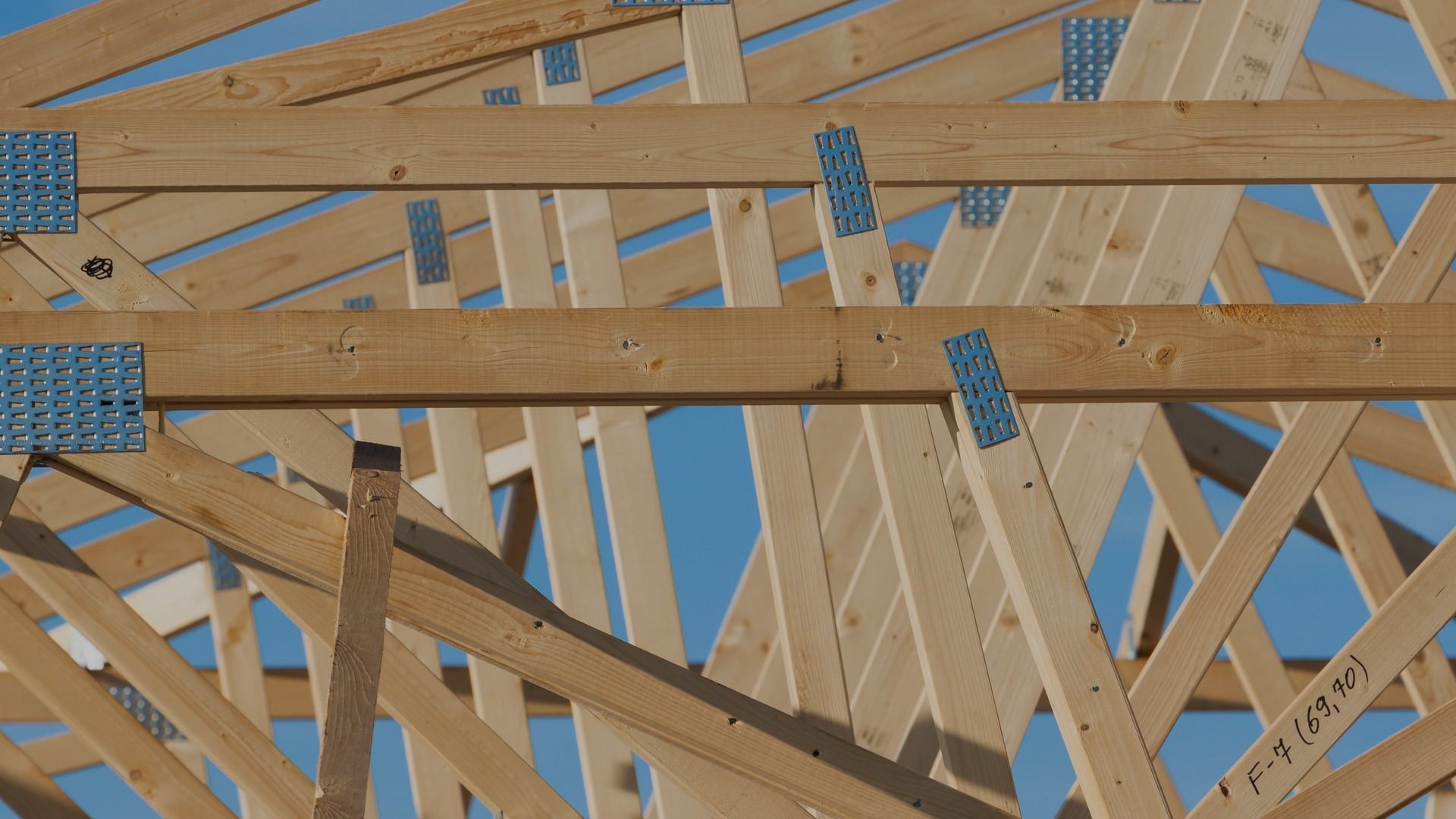 A close-up of wooden roof trusses under construction, showing structural beams connected by metal mending plates.