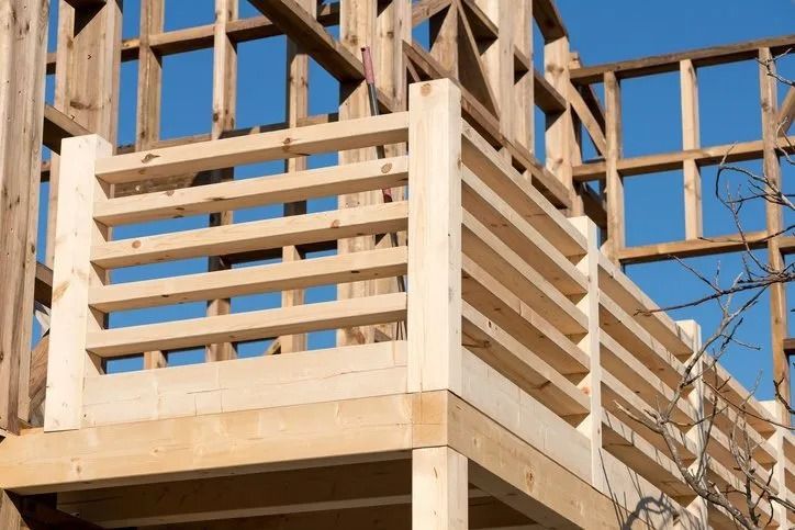 A balcony railing under construction, featuring horizontal wooden planks against a timber-framed building under a blue sky.