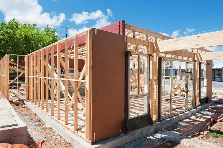 Wooden wall framing and structural beams for a house under construction on a concrete foundation under a sunny sky.