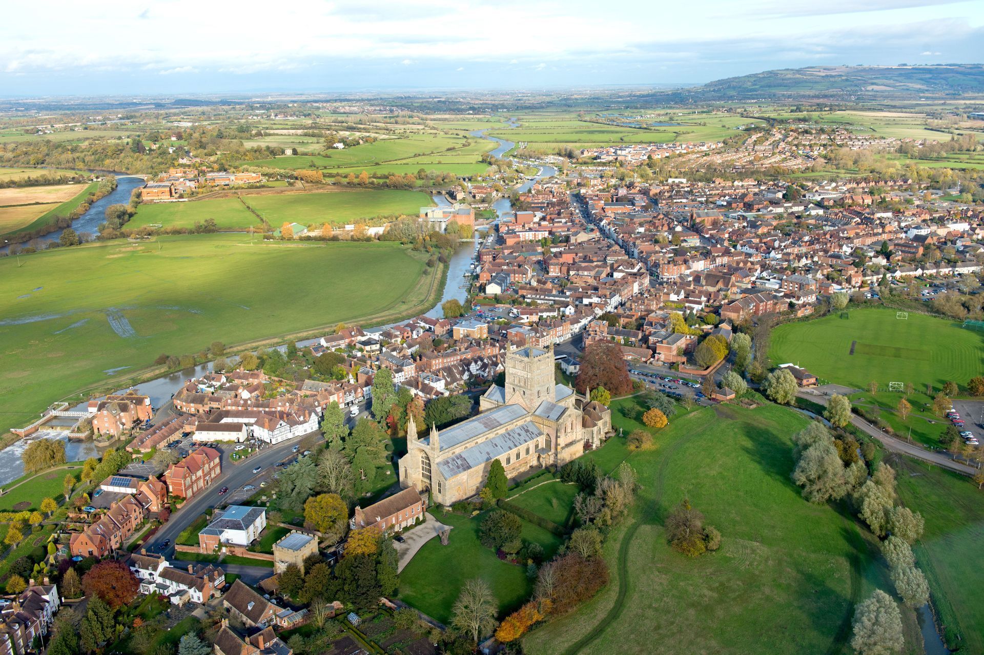 Aerial view of a town with a church, river, green fields, and buildings. Sunny day with mostly brown and green tones.