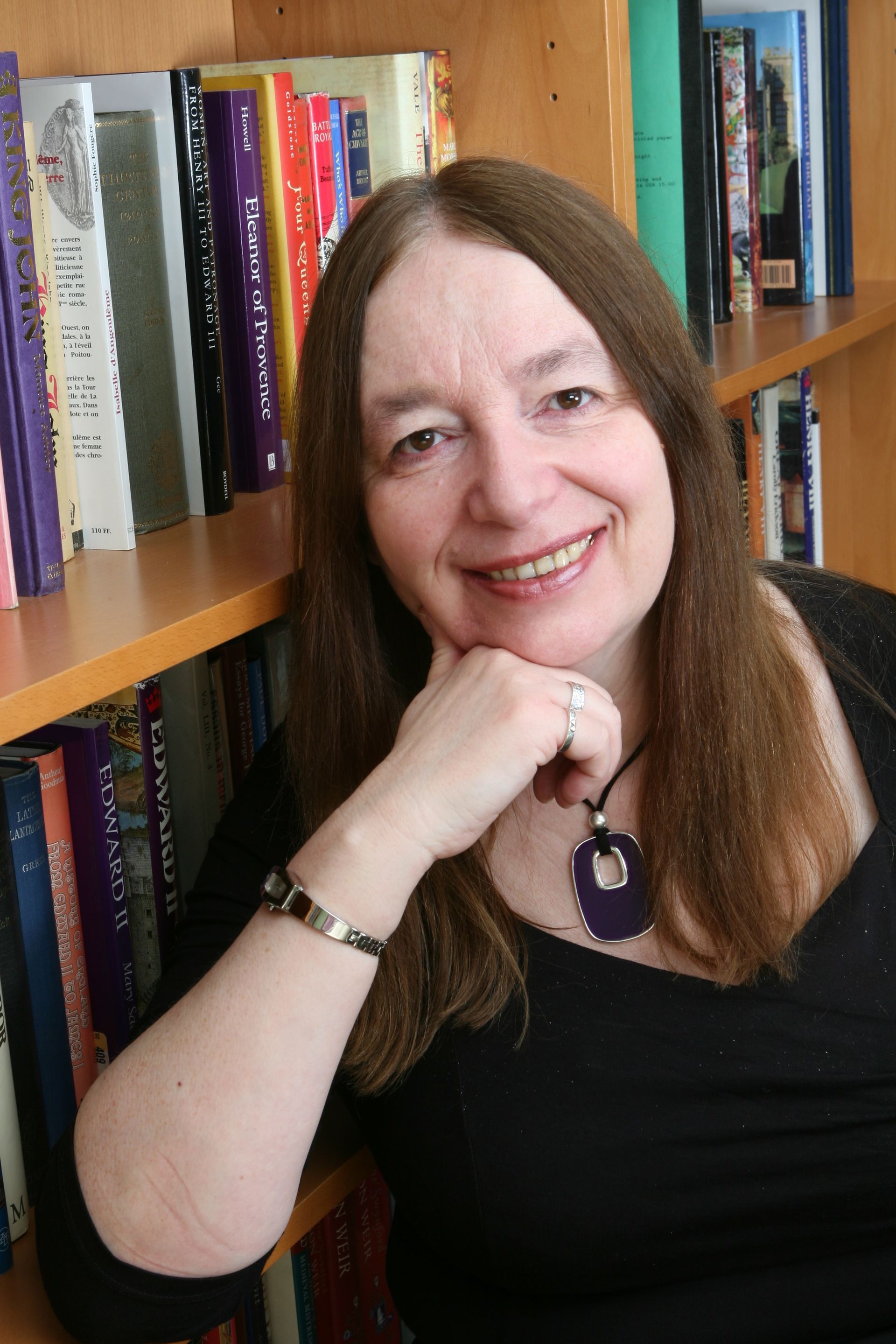 Woman with long brown hair, smiling, leaning on a bookshelf, wearing black top and a pendant necklace.
