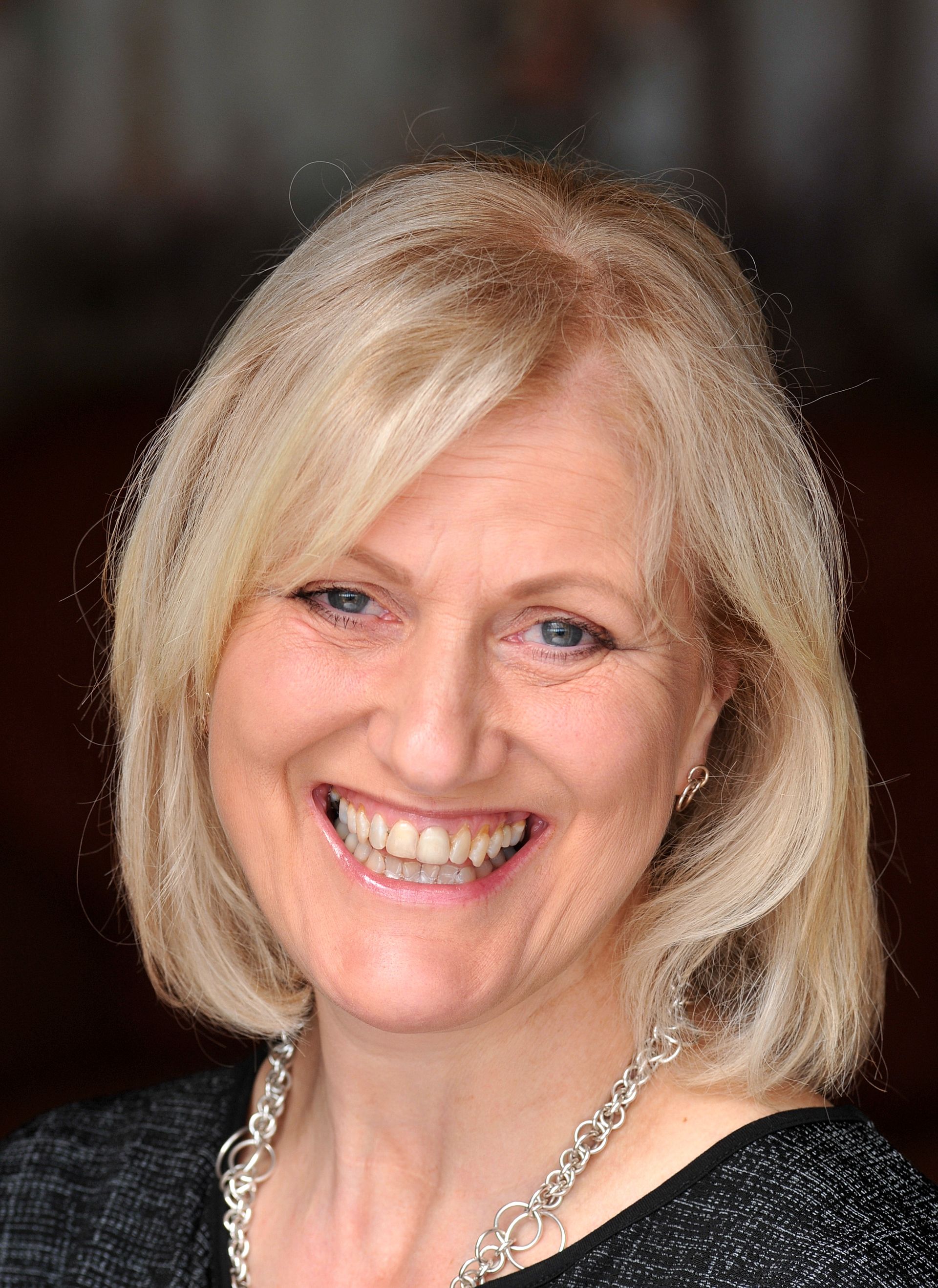 Woman with blonde hair smiling widely, wearing a black top and silver necklace.