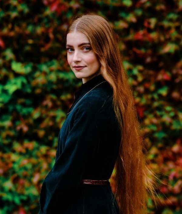 Woman with long red hair, wearing a dark green dress, standing outdoors in front of colorful foliage.