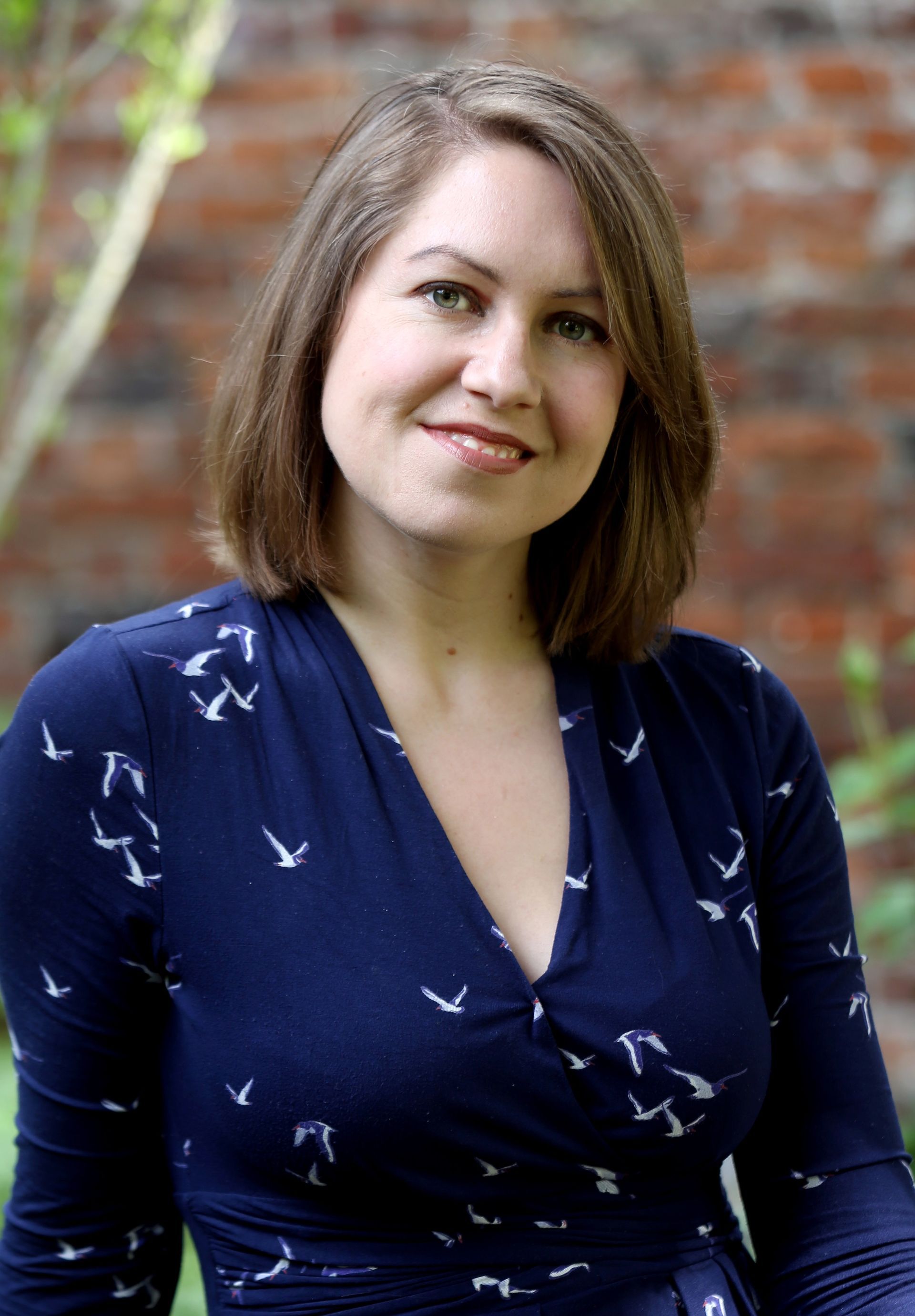 Woman with brown hair in a blue dress, smiling, in front of a brick wall and greenery.