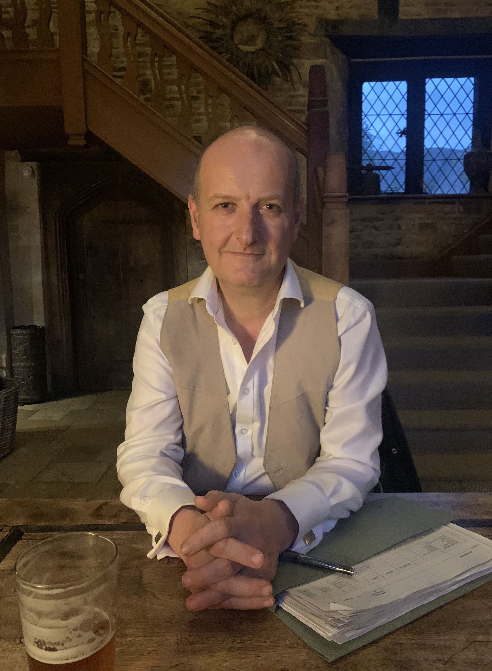 Man in beige vest, white shirt, seated at a wooden table, hands clasped. Interior with stairs, window, and low lighting.