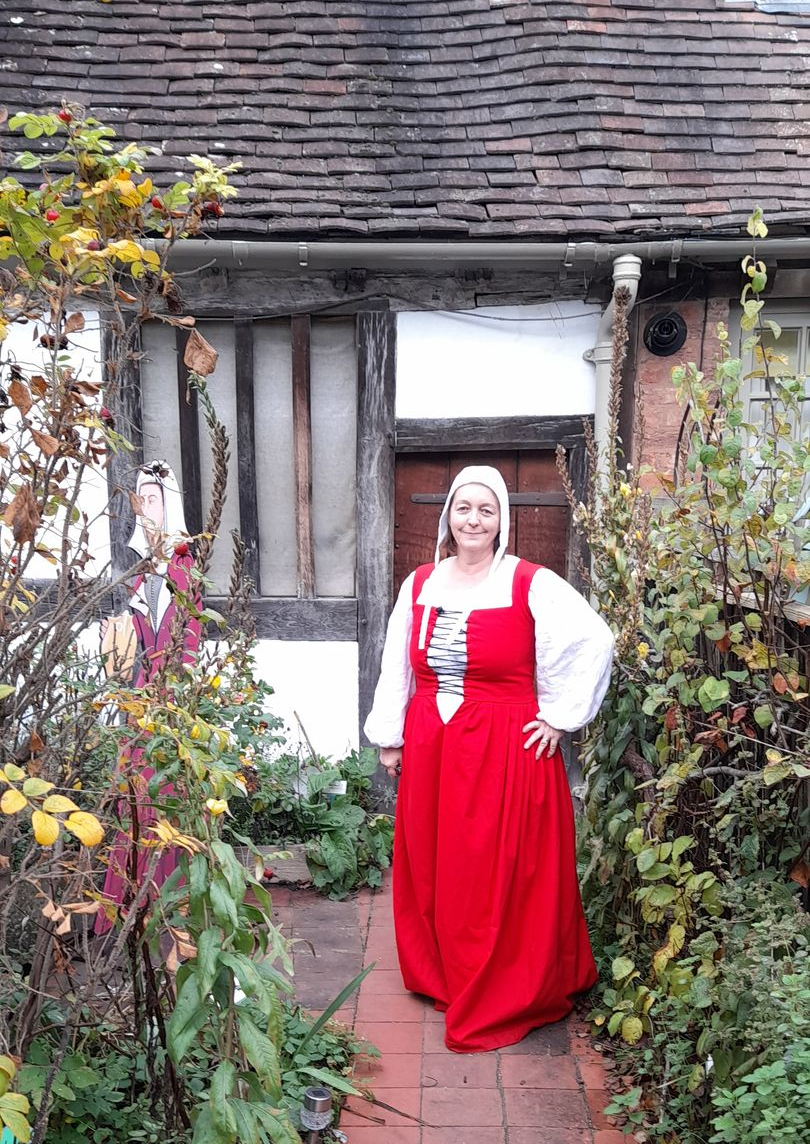 Woman in red medieval-style dress, posing in front of a Tudor-style cottage with timber-framed walls and overgrown garden.