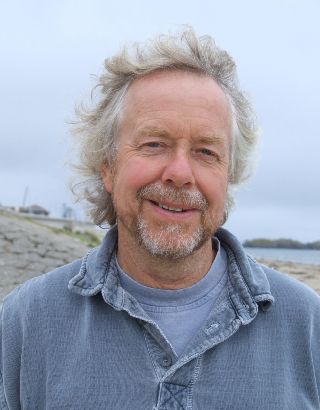 Man with gray hair and beard smiles, outdoors in a blue shirt.