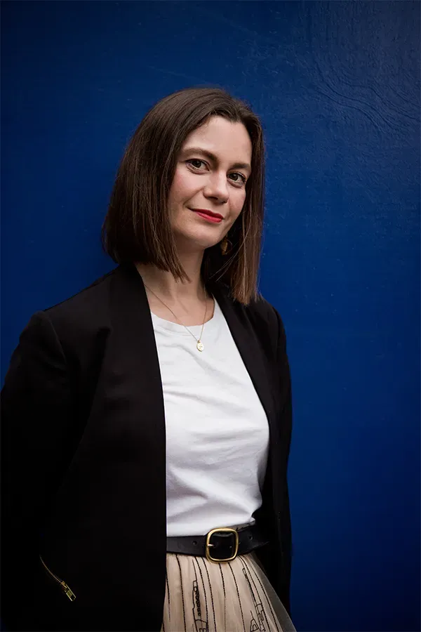 Woman wearing a blazer, white shirt, and skirt, standing against a blue wall.