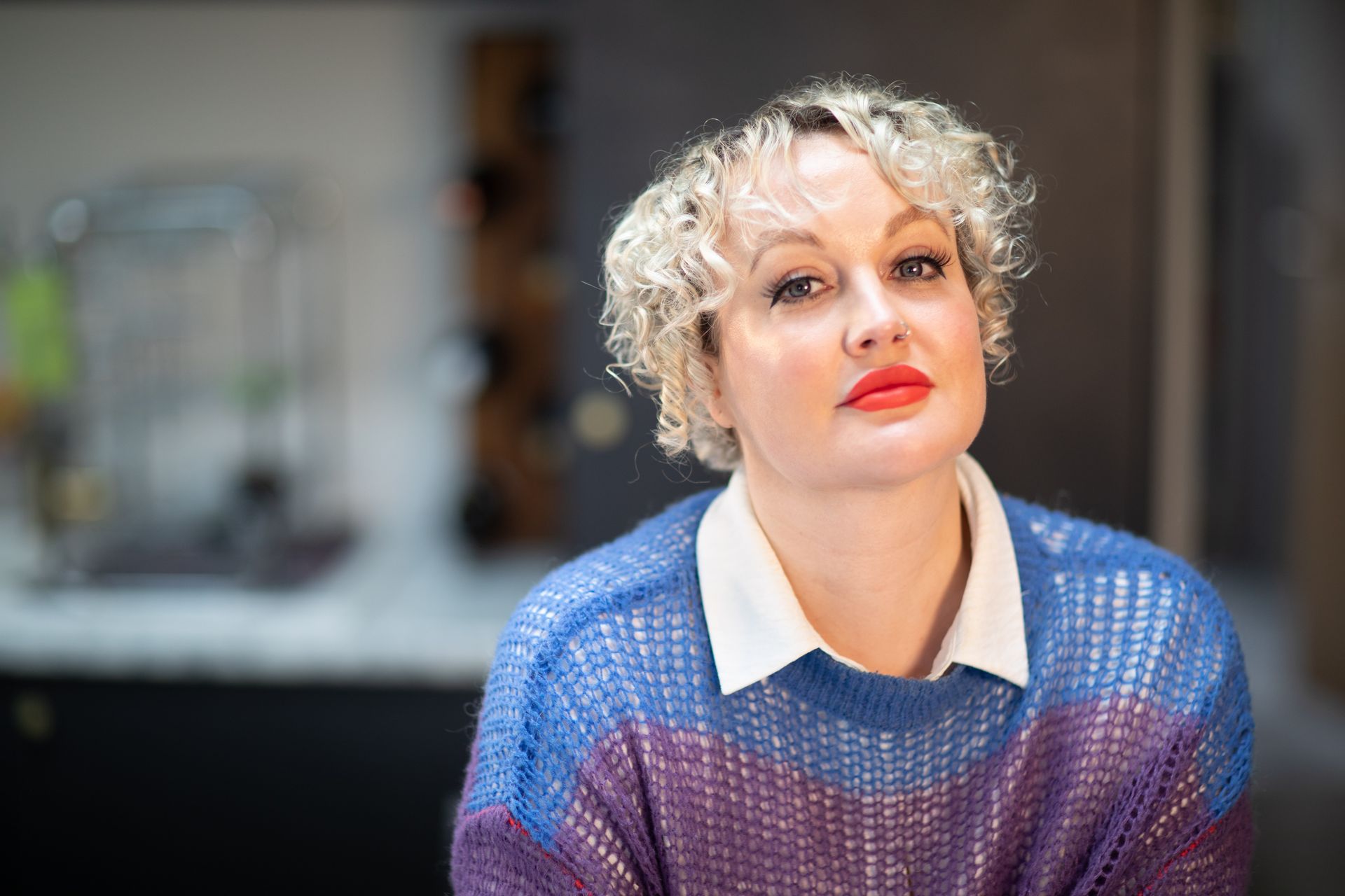 Woman with blonde curly hair wearing a blue and purple sweater and red lipstick, looking towards the camera.