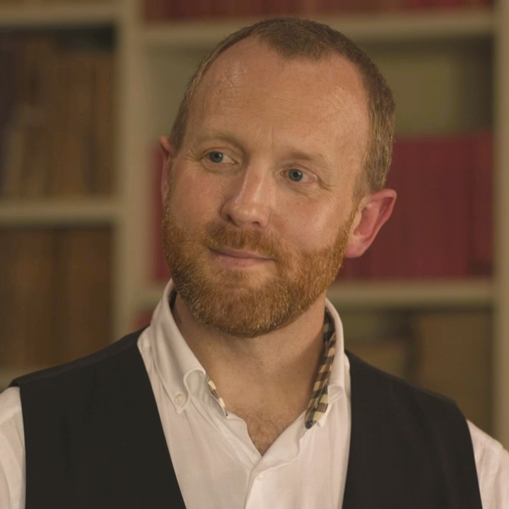 Man with ginger beard, wearing a vest and white shirt, smiling slightly, looking off-camera.