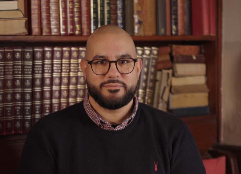 Bald man with glasses and a beard in front of a bookshelf, wearing a black sweater and patterned shirt collar.