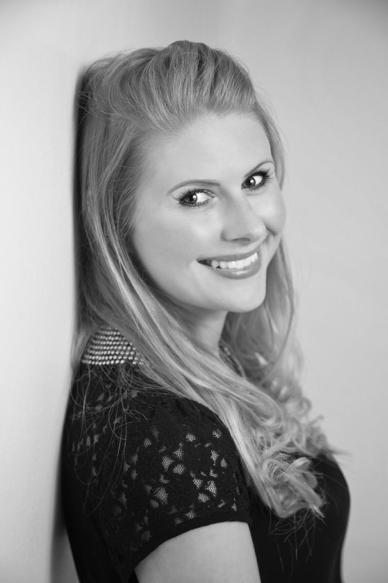 Woman with blonde hair smiles, leaning against a wall. Black lace top, formal setting.