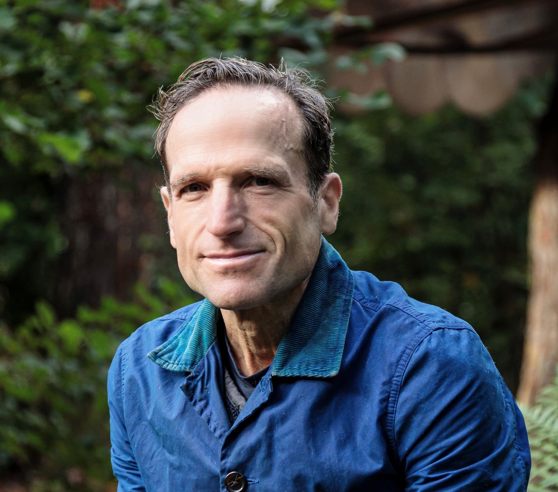 Man in blue jacket with teal collar smiles outdoors, surrounded by foliage.