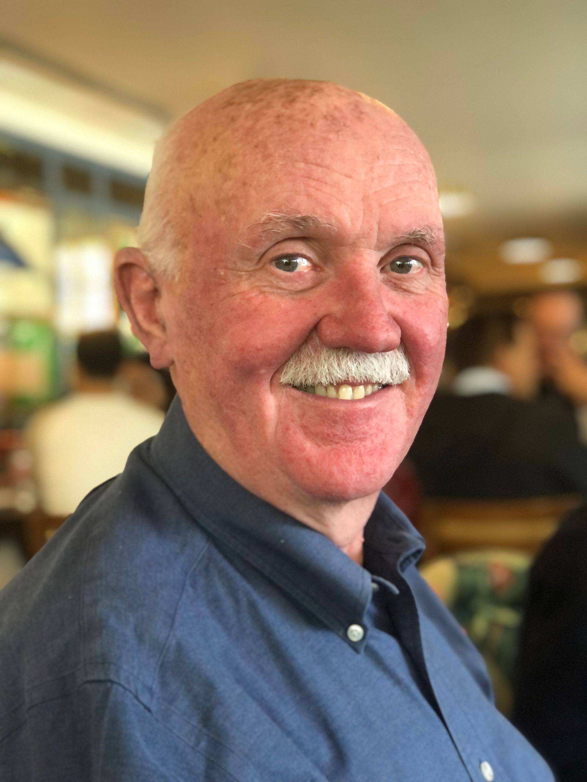 Smiling man with a white mustache wearing a blue shirt, seated indoors.