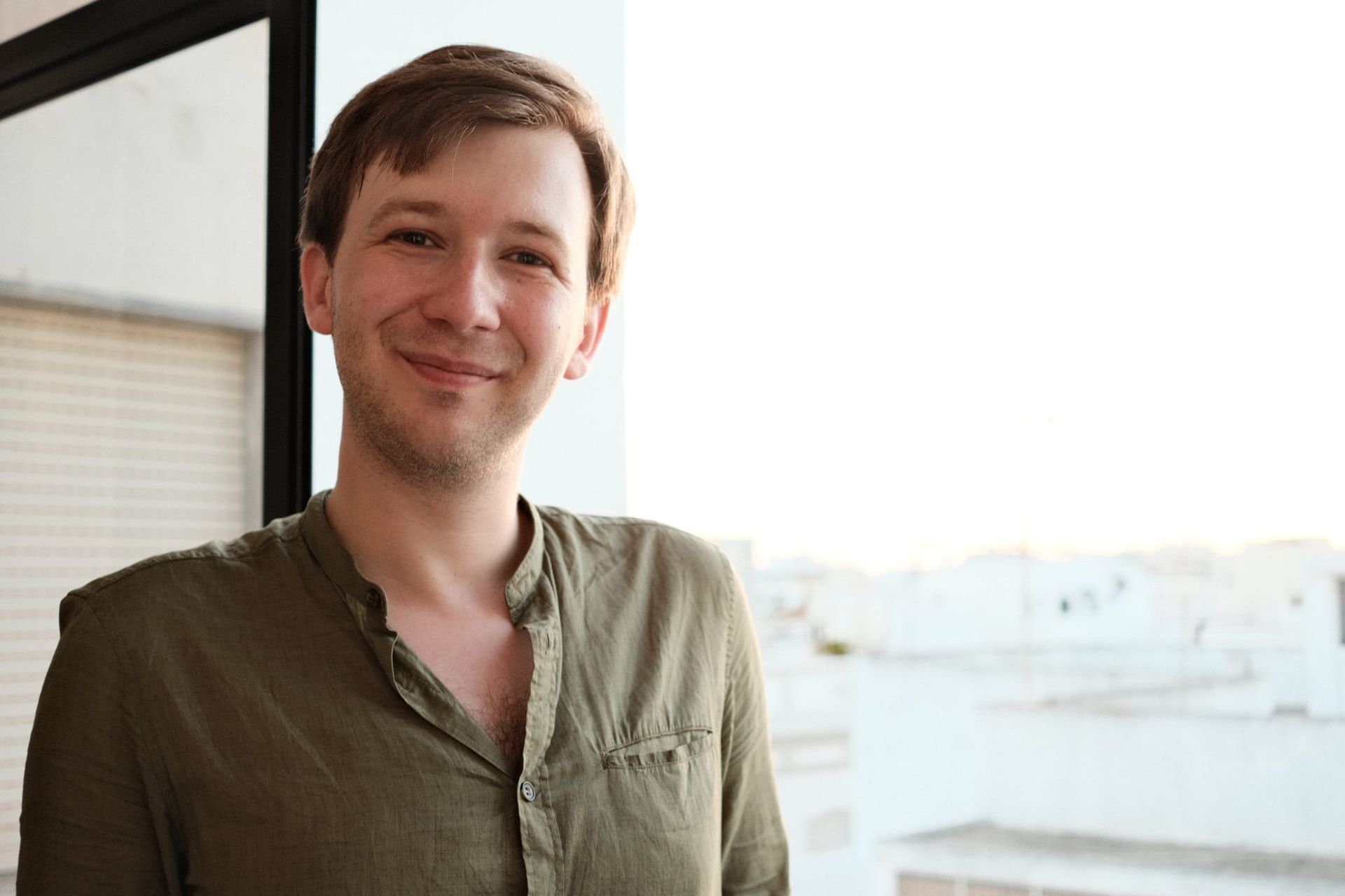 Man in green shirt smiles by a window, overlooking a bright outdoor scene.
