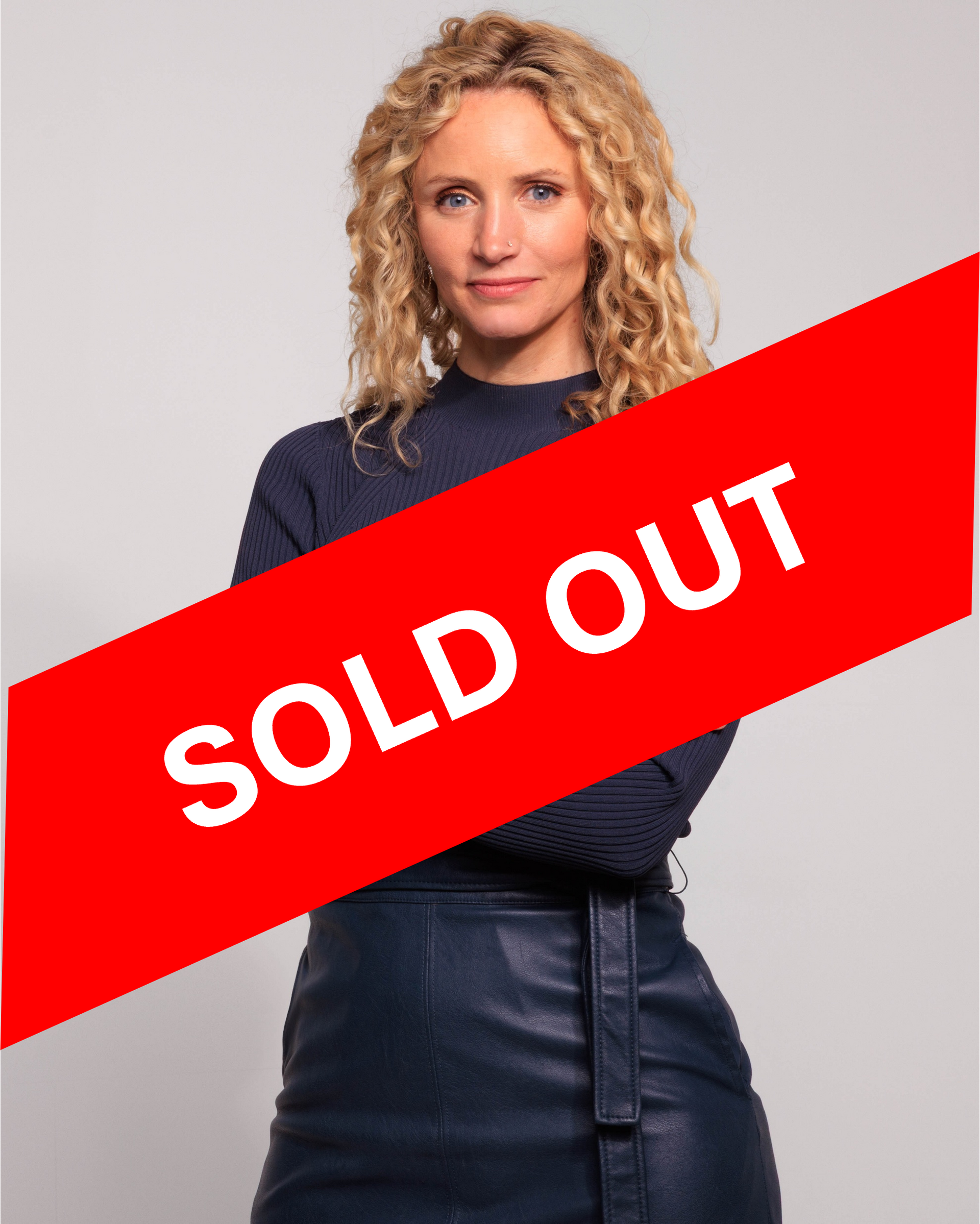 Woman with curly blonde hair, arms crossed, wearing a navy blue sweater and dark blue skirt.