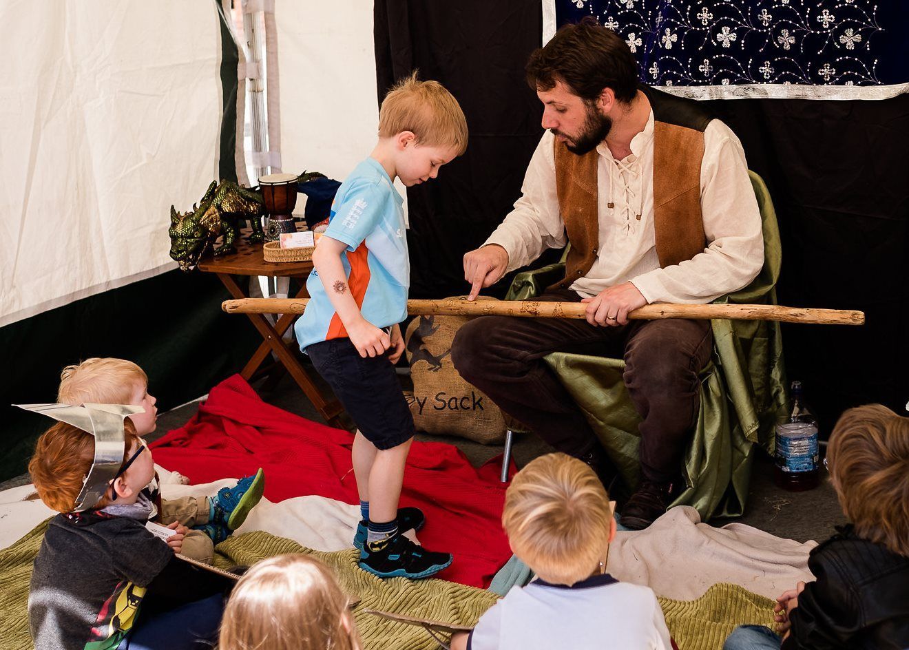 Man showing a stick to a child, surrounded by other children. Indoors, tent-like setting.