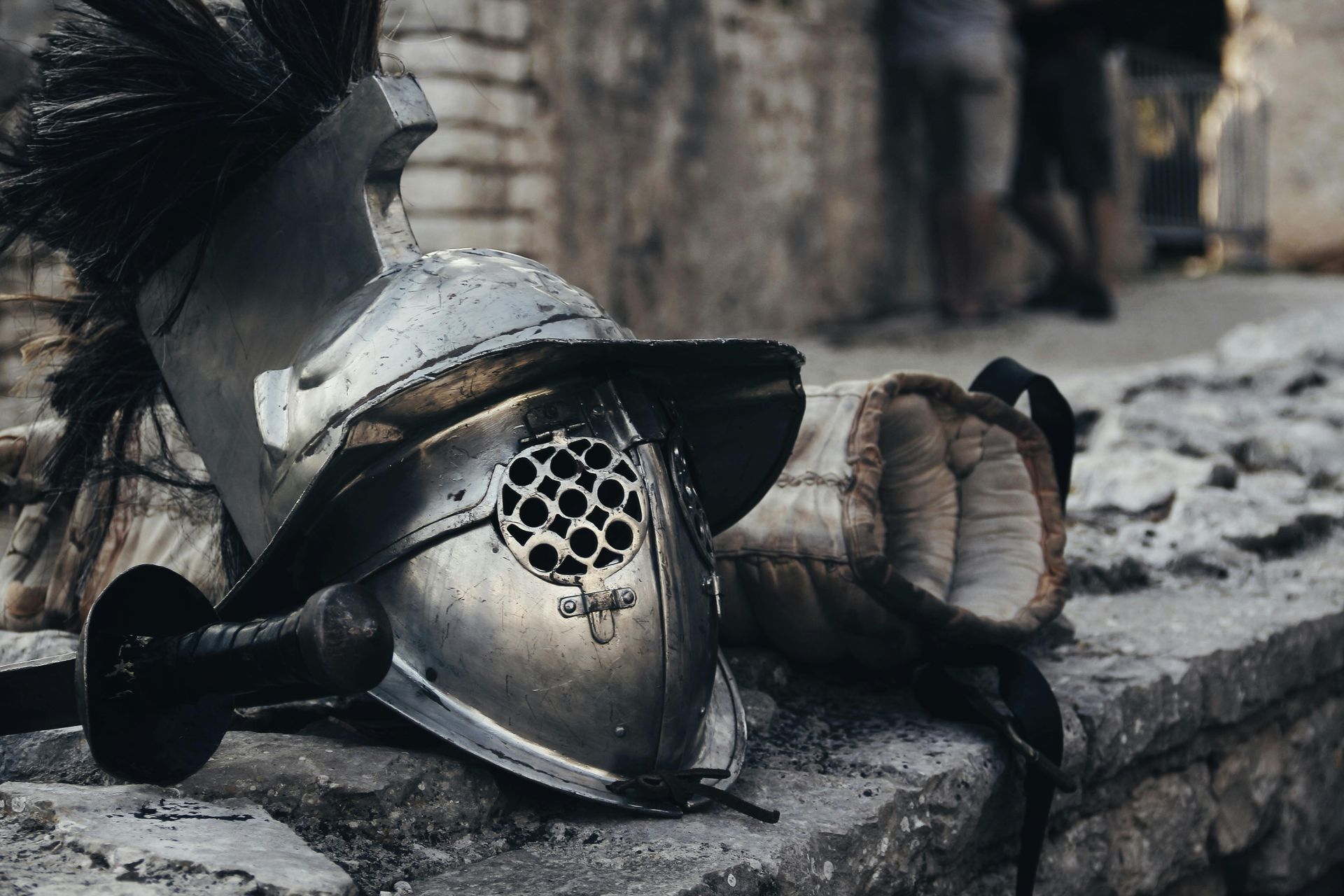 A gladiatorial helmet and arm guard rest on a stone surface, set outdoors.