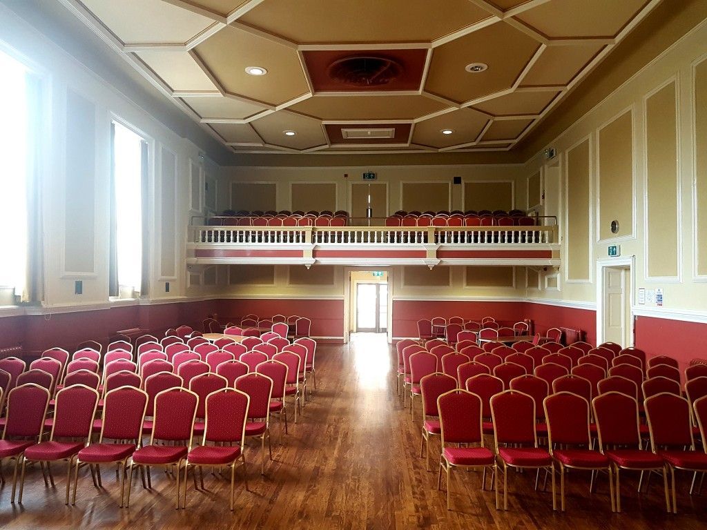 Rows of red chairs face a stage in a large hall with a balcony and ornate ceiling.