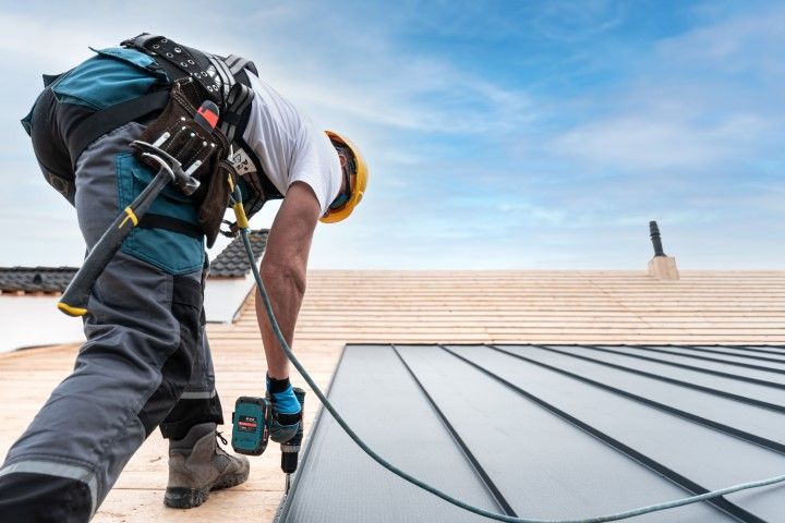 Worker installing metal roofing on a rooftop, carrying tools and wearing a harness