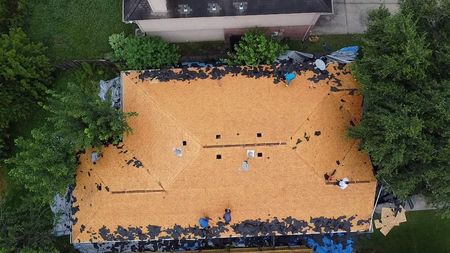 An aerial view of a house roof undergoing renovation with workers removing old shingles to expose the wooden deck.
