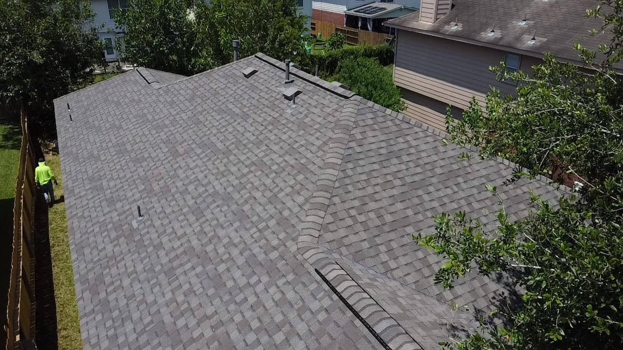 A high-angle view of a grey shingled roof on a sunny day, with a person in a yellow safety vest visible on the ground.