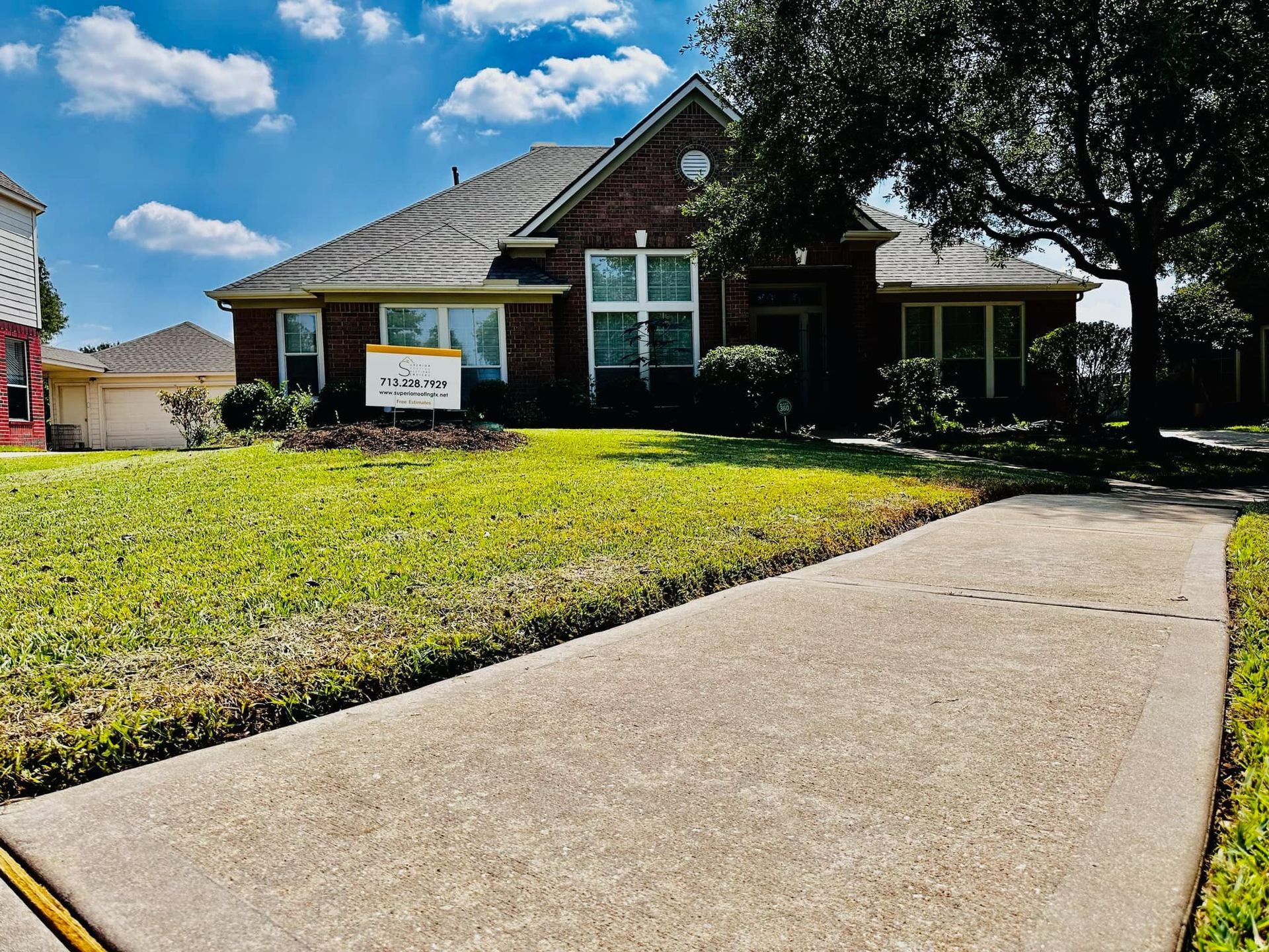 A brick, single-story house with a lawn, a concrete walkway in the foreground, and a business sign on a sunny day.