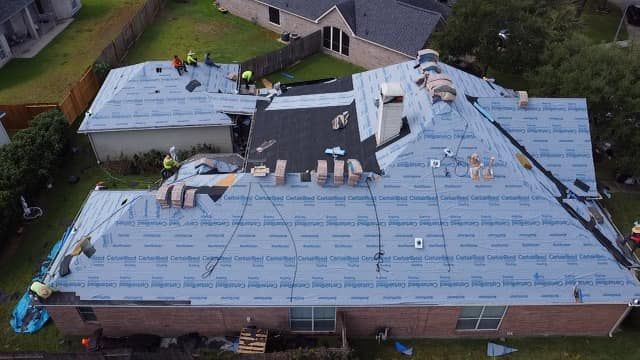 Aerial view of a house roof undergoing repairs, covered in blue underlayment paper, with construction workers present.