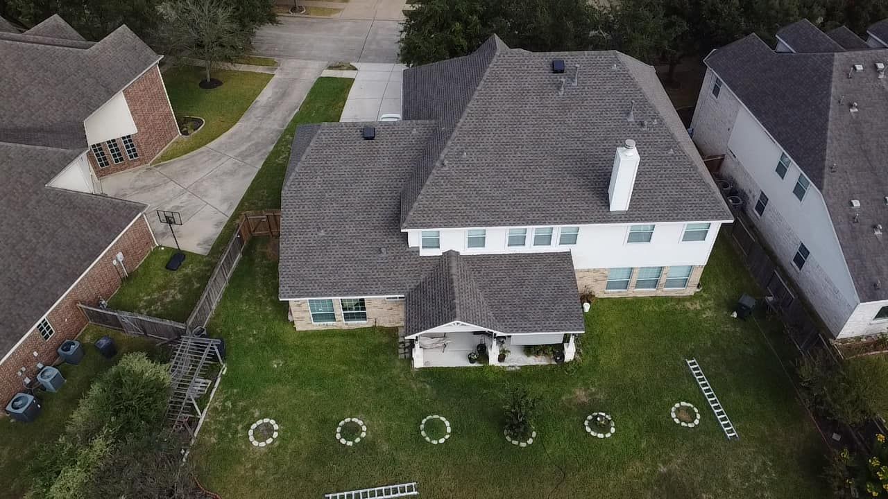 An aerial view of a two-story suburban house with a gray shingled roof, a chimney, and a grassy backyard.