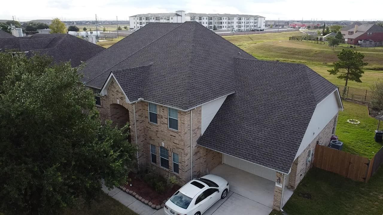 An aerial view of a two-story brick suburban house with a dark shingled roof, a front driveway, and a white parked car.