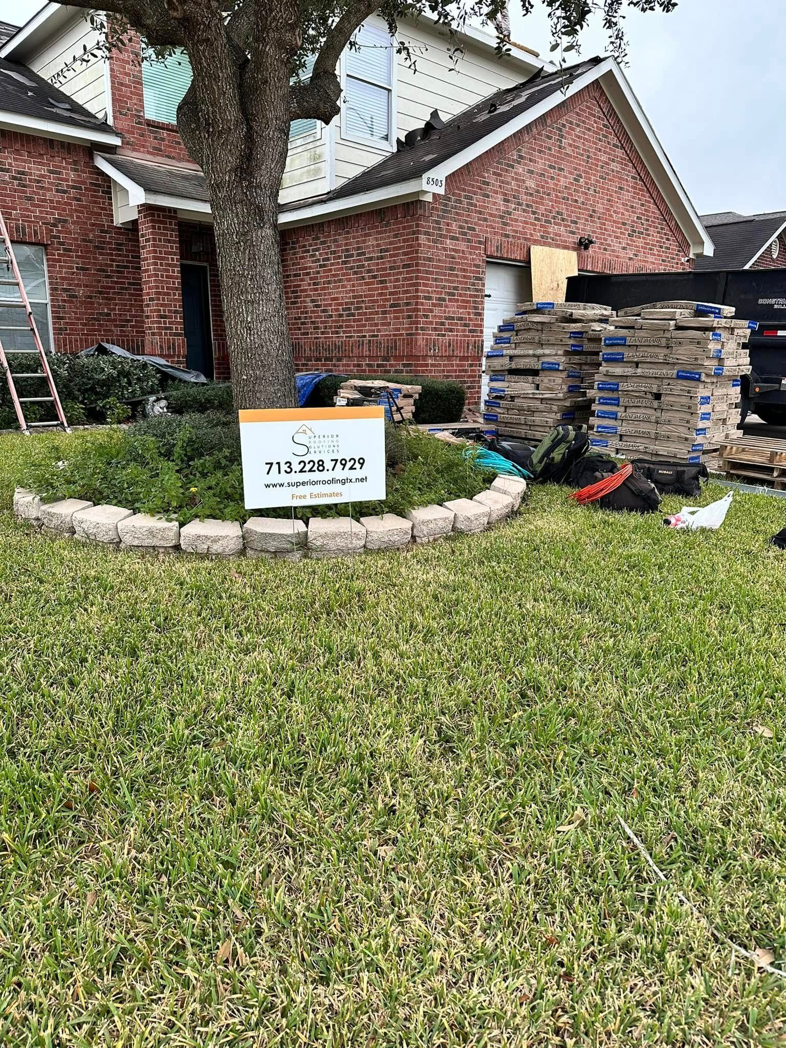 A brick house under renovation with a yard sign, a tree-shaded garden bed, and a stack of construction materials nearby.