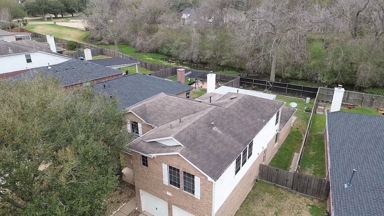 An aerial view of suburban brick townhouses with dark shingled roofs and white chimneys, situated near a wooded area.