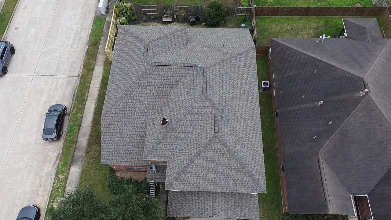 Aerial view of a gray shingled residential roof with several vents, located next to a street with parked cars.