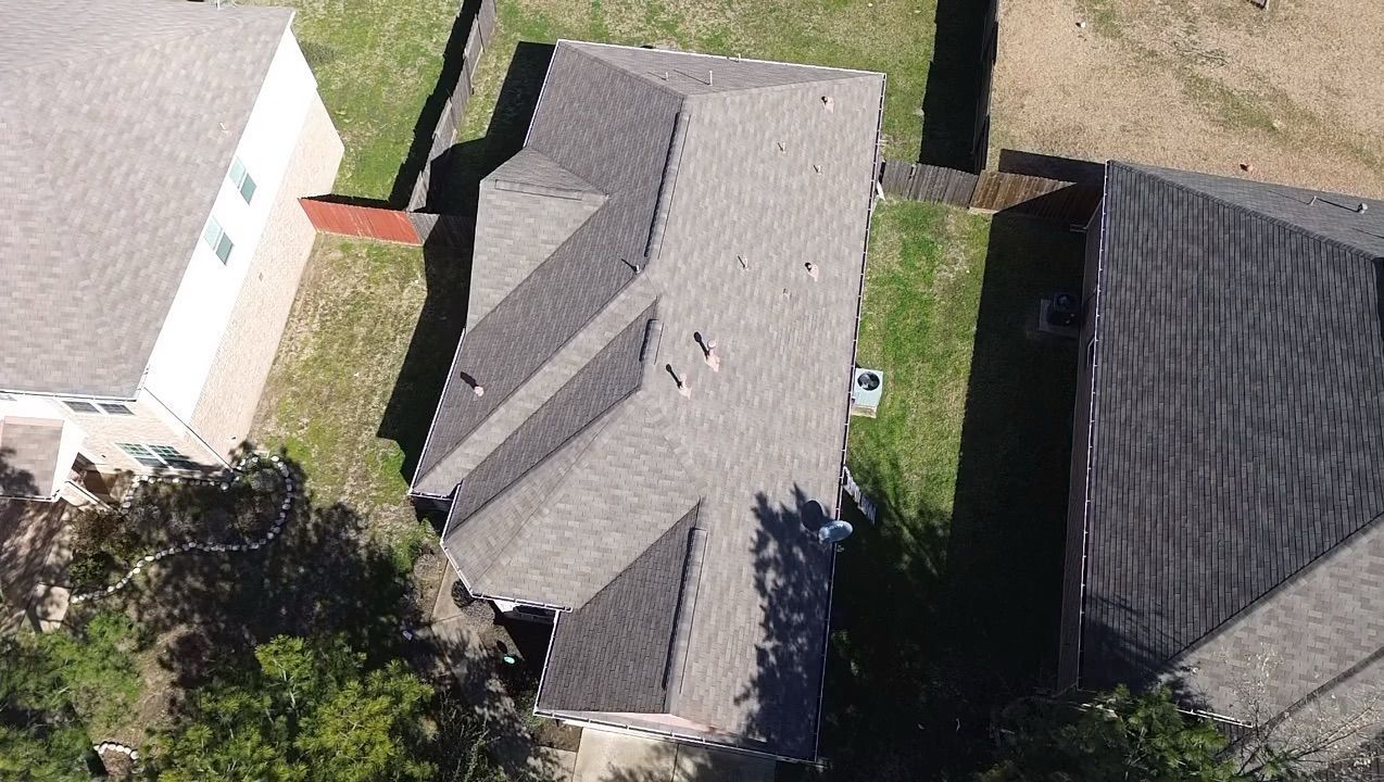 An aerial view of a gray shingled residential house roof, surrounded by green lawns and neighboring homes.