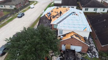 Workers remove old shingles and install new underlayment on a residential roof in a suburban neighborhood.